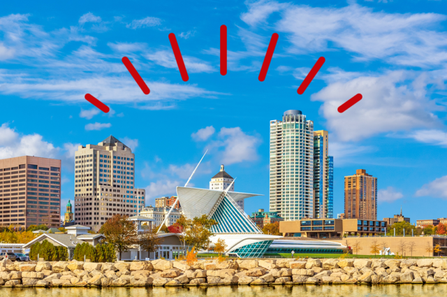 The Milwaukee downtown skyline set against a blue sky is reflected in the waters of Lake Michigan in the foreground.